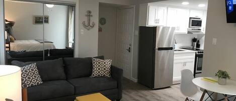 A modern living room with a sofa, ottoman, and a small table. A kitchen is visible in the background with a refrigerator and a microwave.