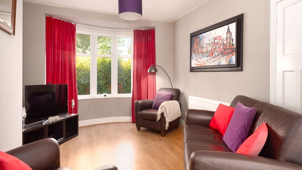 Een moderne woonkamer met een flatscreen televisie, leren banken, rode gordijnen en een schilderij van een stadsgezicht.