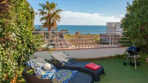 Eine Terrasse mit zwei Lounge-Stühlen, einem Grill und Blick aufs Meer.