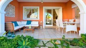 Un'area salotto esterna accogliente con divano, sedie e tavolo, immersa in un giardino con piante e rocce.