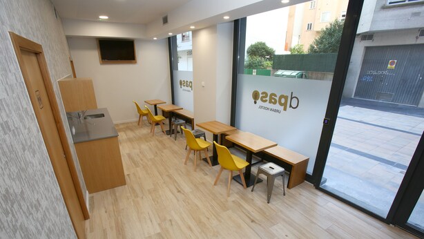 A modern cafe with wooden tables and yellow chairs, a sink, and a large window with a view of the street. - dpaso Urban Hostel (Pontevedra)