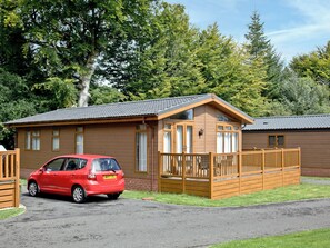 A single-story wooden cabin with a fenced deck, a car parked in front, and a natural backdrop of trees. - 2 bedroom accommodation in Lanlivery (Lanlivery)