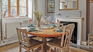 Une salle à manger avec une table en bois dressée pour quatre personnes, entourée de chaises. On y trouve une cheminée avec un manteau de cheminée, une fenêtre avec des rideaux et un lustre.