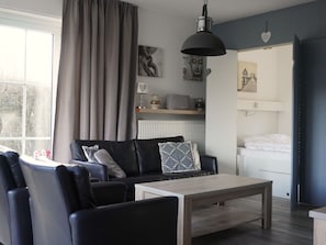 A modern living room with a sofa, a coffee table, and a dining table. There is a window with curtains, a radiator, and wall art.