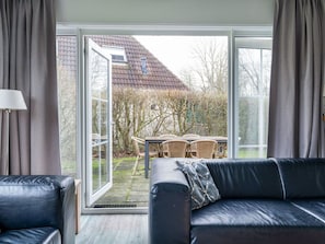 A modern living room with a black leather sofa, a table with chairs, and a large sliding glass door leading to an outdoor area with a patio and a view of a house.