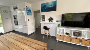 A modern kitchen with a built-in oven and microwave, a dining area with a wooden table, and a living area with a TV and decorative items. - Villa Sandy Beach - Capbreton (Capbreton)