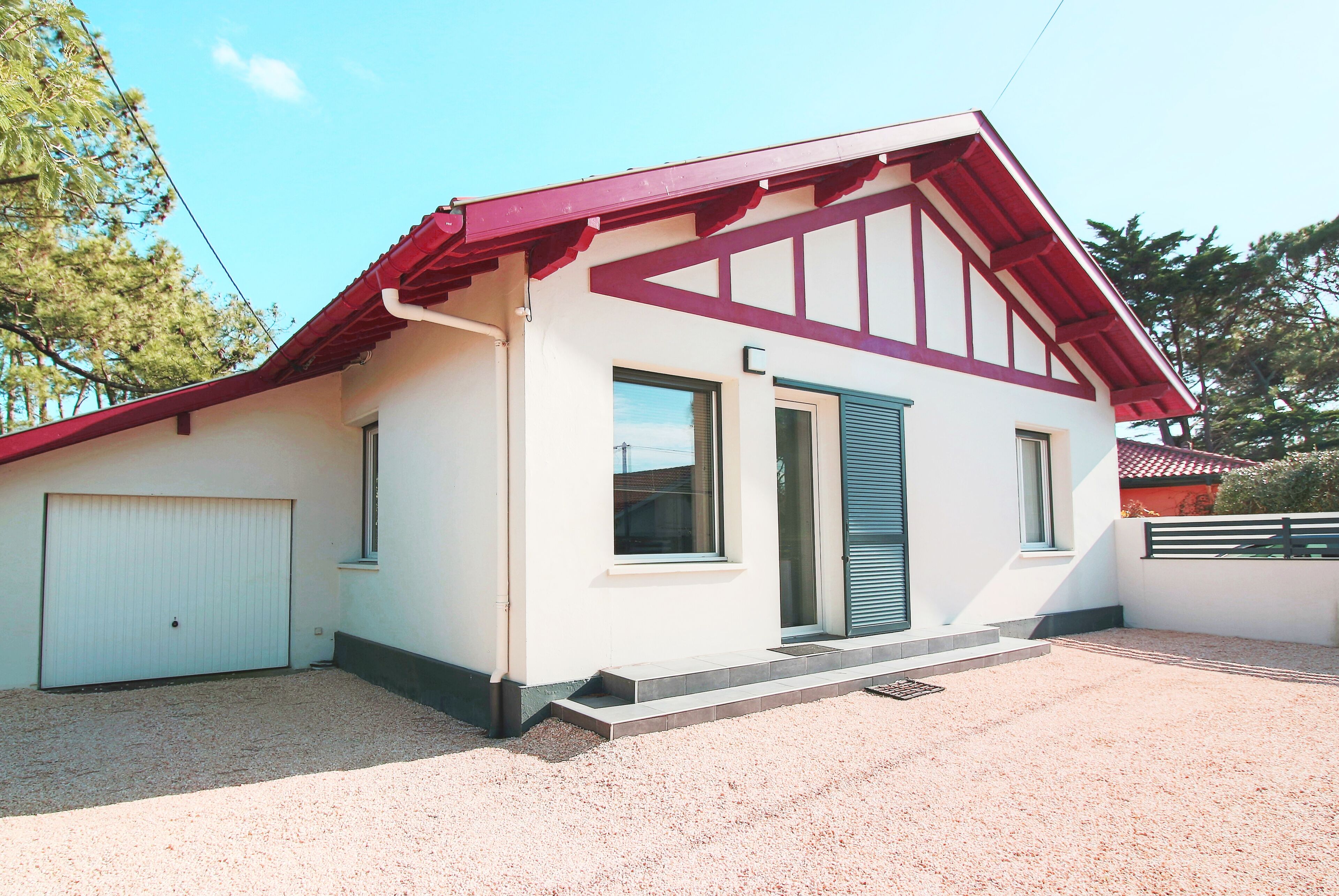 Ein eingeschossiges Haus mit rotem Dach und weißen Wänden. Es besitzt eine Garagentür, ein kleines Fenster und eine Tür mit grünem Fensterladen.
