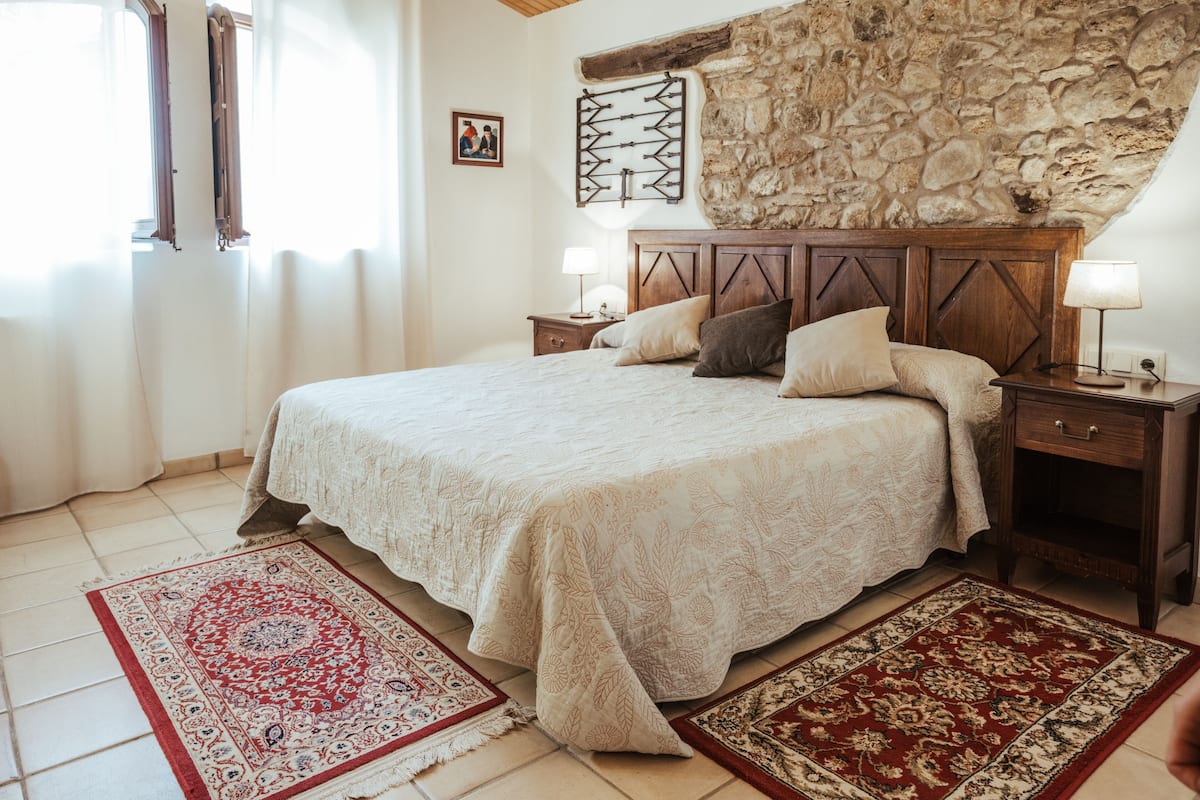 Un dormitorio con pared de piedra, estructura de cama de madera, dos mesitas de noche con lámparas y una alfombra estampada en rojo y blanco.