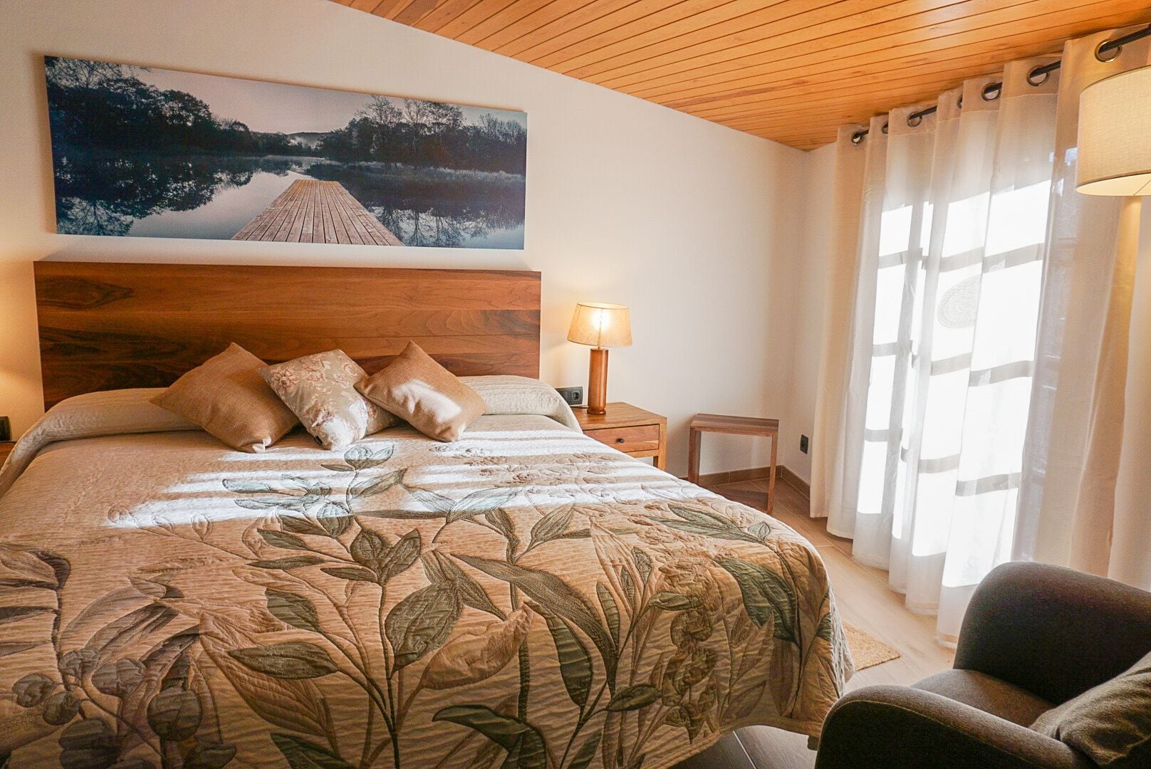 A bedroom with a wooden headboard, a bed with floral bedding, a bedside table with a lamp, and a view of a lake through sheer curtains.