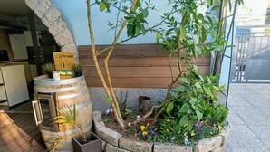 Een rustieke buitenruimte met een houten bankje, een trommelvormige plantpot met bloemen en een kleine tafel met een bord.