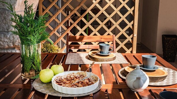 Een houten tafel gedekt met een witte taart, twee appels, een glazen vaas met groen en een houten stoel.