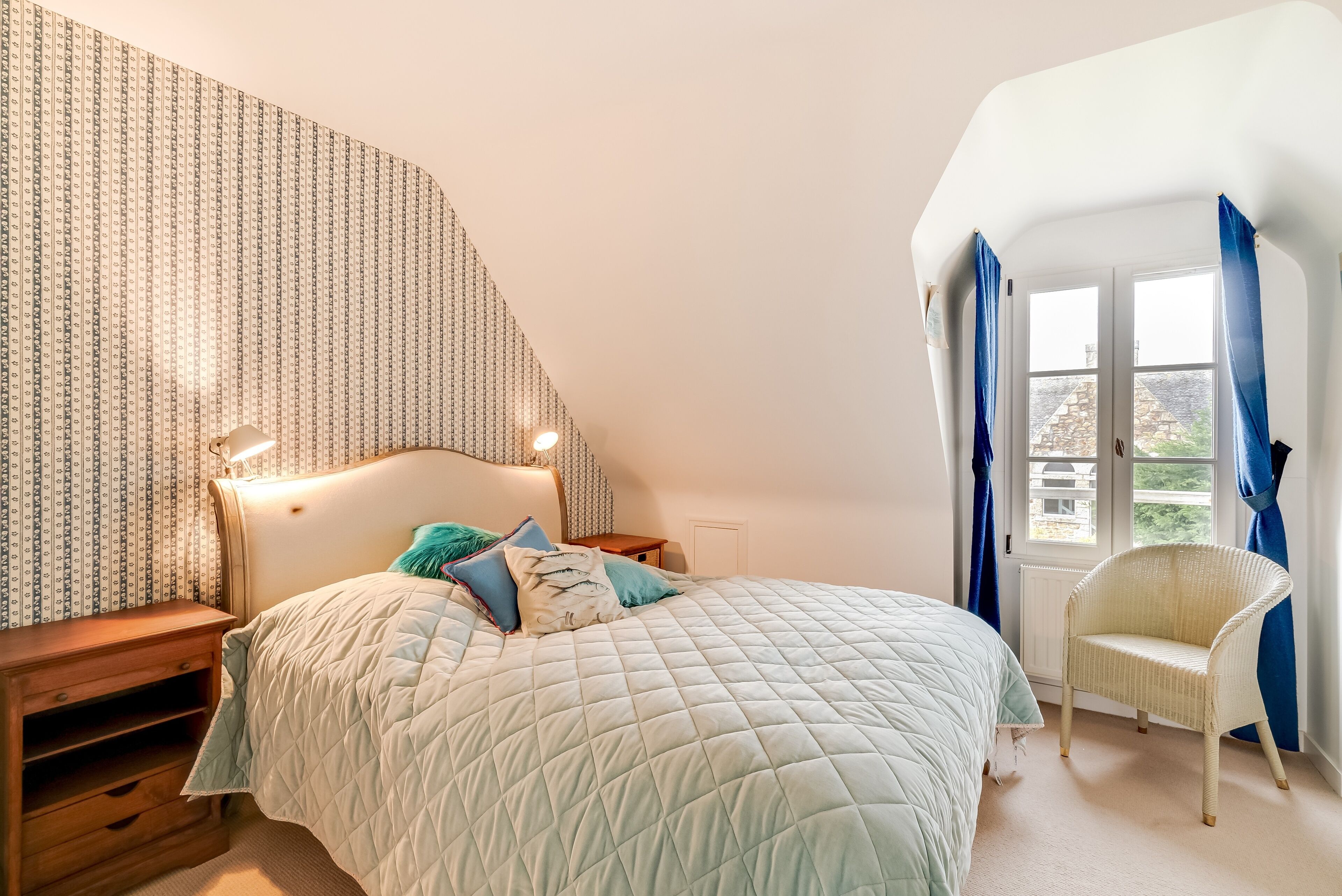 A bedroom with a large bed, a nightstand, a chair, and a window with curtains.