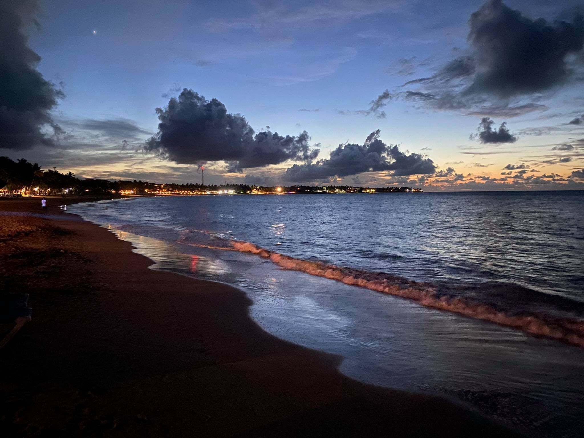 Una spiaggia con le onde al tramonto, una linea di costa cittadina in lontananza e un cielo nuvoloso.