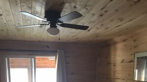 Ein Schlafzimmer mit einem Bett, Deckenventilator, einem Fenster mit Vorhängen und Blick ins Freie.