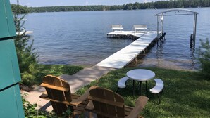 Eine Seeterrasse mit Holzstühlen und einem Tisch, eine Anlegestelle, die ins Wasser führt, und ein bewaldeter Uferstreifen im Hintergrund.