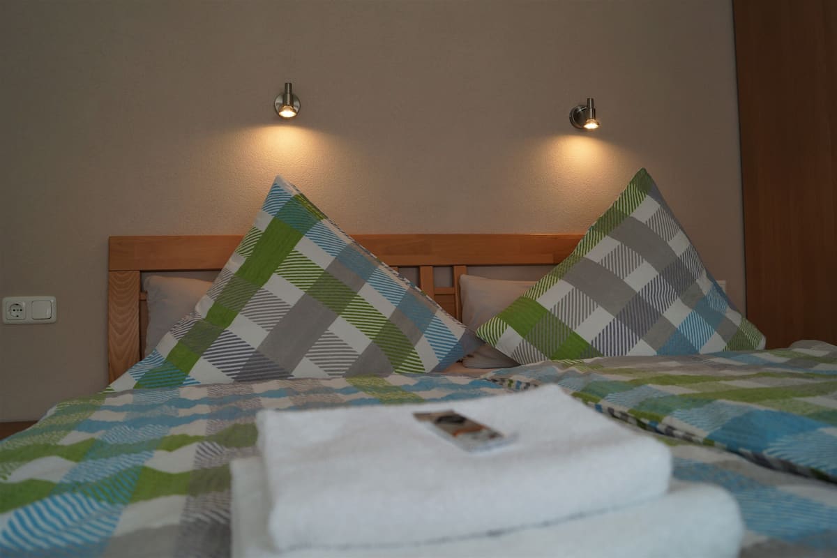 Habitación de hotel con cabecera de madera, dos almohadas con un diseño geométrico y una toalla blanca doblada con cuidado sobre la cama.