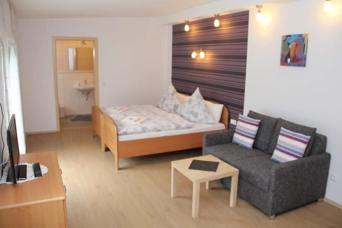 Un dormitorio moderno con cama, sofá gris, una mesita y un escritorio con una computadora portátil.