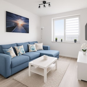 Una sala moderna con un sofá azul, una mesa de centro blanca, un cuadro de un atardecer sobre el agua, una lámpara de pie y una ventana con persianas.