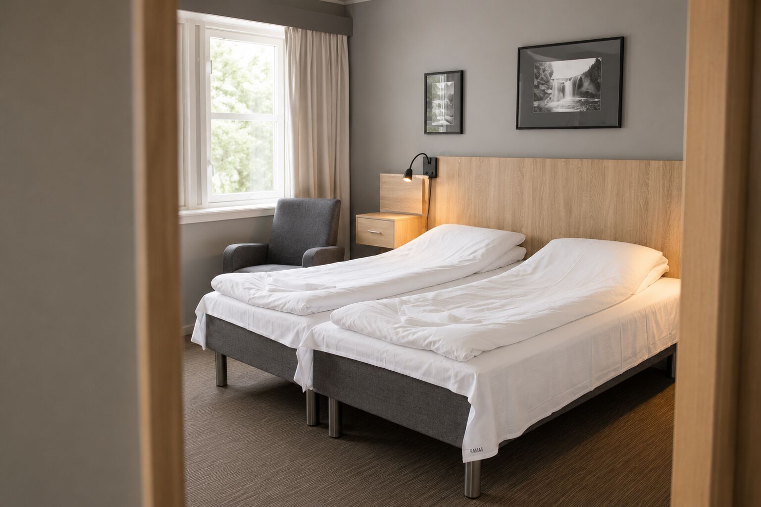 Habitación de hotel con dos camas, una silla, una mesita de noche, una lámpara y dos cuadros en la pared.
