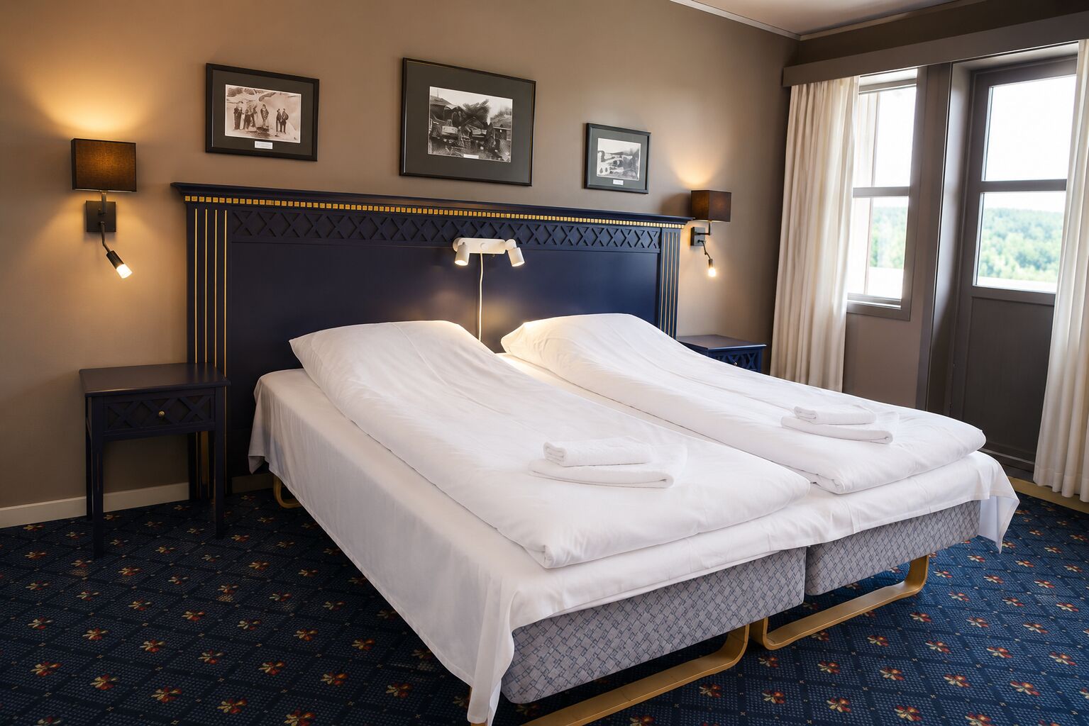A double bed with white linens, a dark headboard, wall-mounted lamps, and framed pictures on the wall.