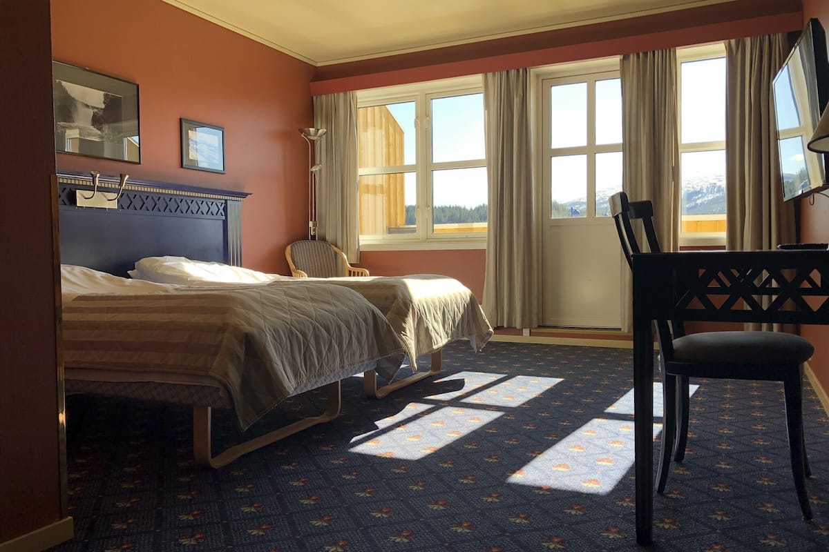 Habitación de hotel con dos camas, una silla, un escritorio y ventanas grandes con cortinas.