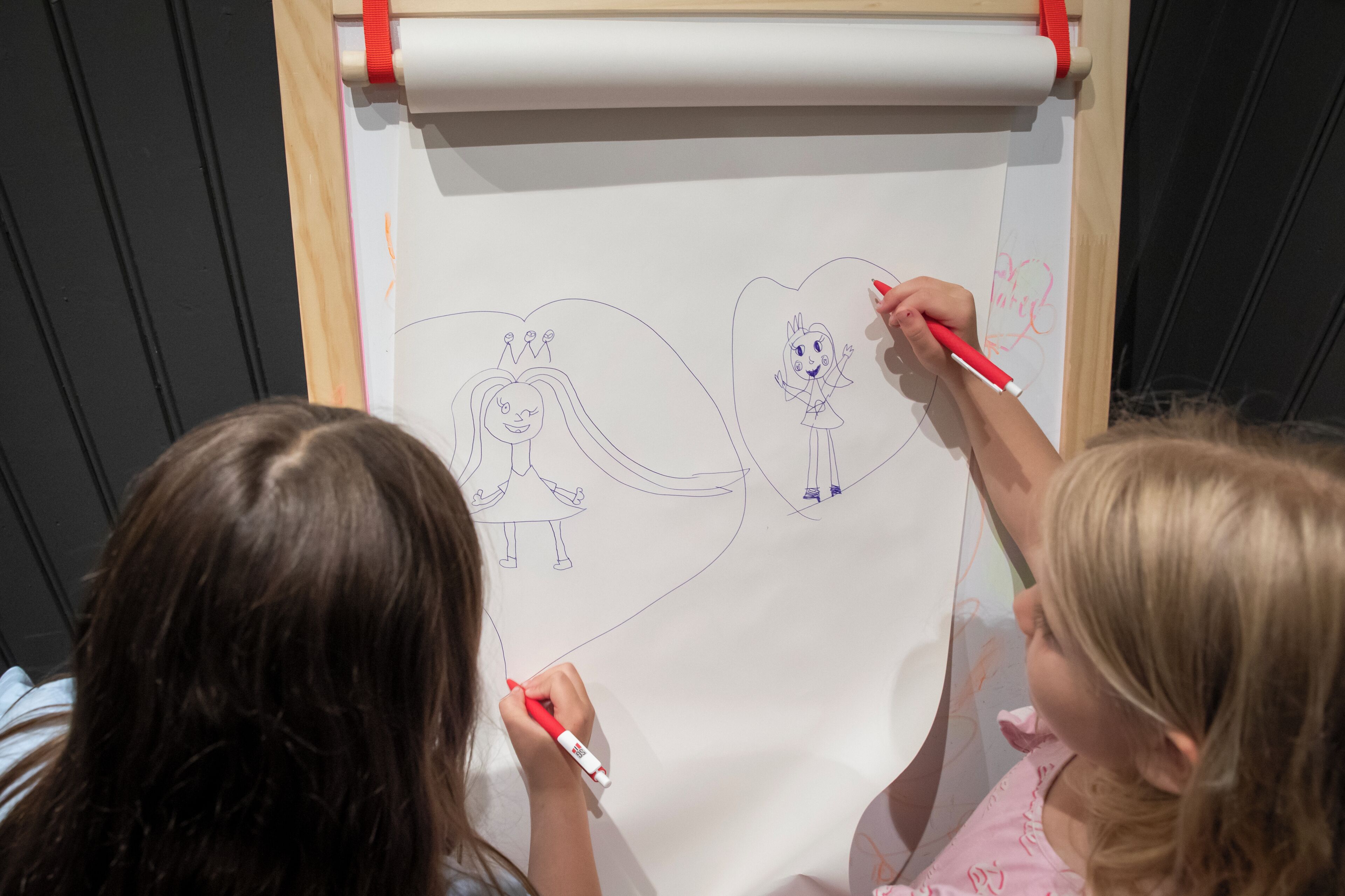 Two individuals drawing on a large white sheet of paper, with a wooden easel and red tape visible.