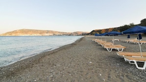 Ein Strand mit Reihen von Liegestühlen und Sonnenschirmen, ein Kiesstrand und ein ruhiges Meer mit Hügeln im Hintergrund.