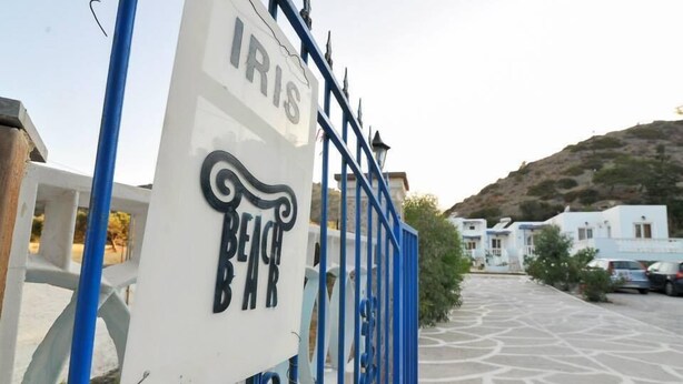 Ein verschlossenes Eingangstor mit einem Schild mit der Aufschrift IRIS BEACH BAR und Blick auf eine Küstenregion mit Gebäuden und geparkten Autos.