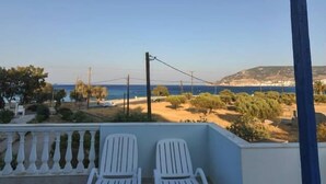 Ein Balkon mit zwei weißen Plastikstühlen, einem beigen Sonnensegel und Blick auf das Meer und die Hügel.