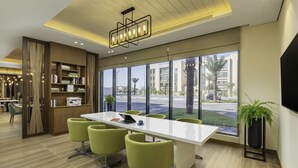 A modern hotel lobby with a long white reception desk, green chairs, a bookshelf, and large windows offering a view of palm trees and buildings. - Holiday Inn & Suites Al Jubail by IHG (Al Jubail)
