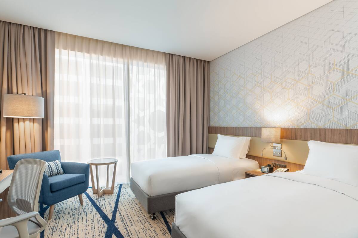 Ett hotellrum med två sängar, ett skrivbord, en stol, ett litet bord och en lampa.