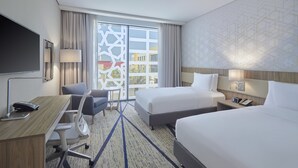 A hotel room with a desk, two beds, a chair, a TV, and a window with a view of buildings. - Holiday Inn & Suites Al Jubail by IHG (Al Jubail)