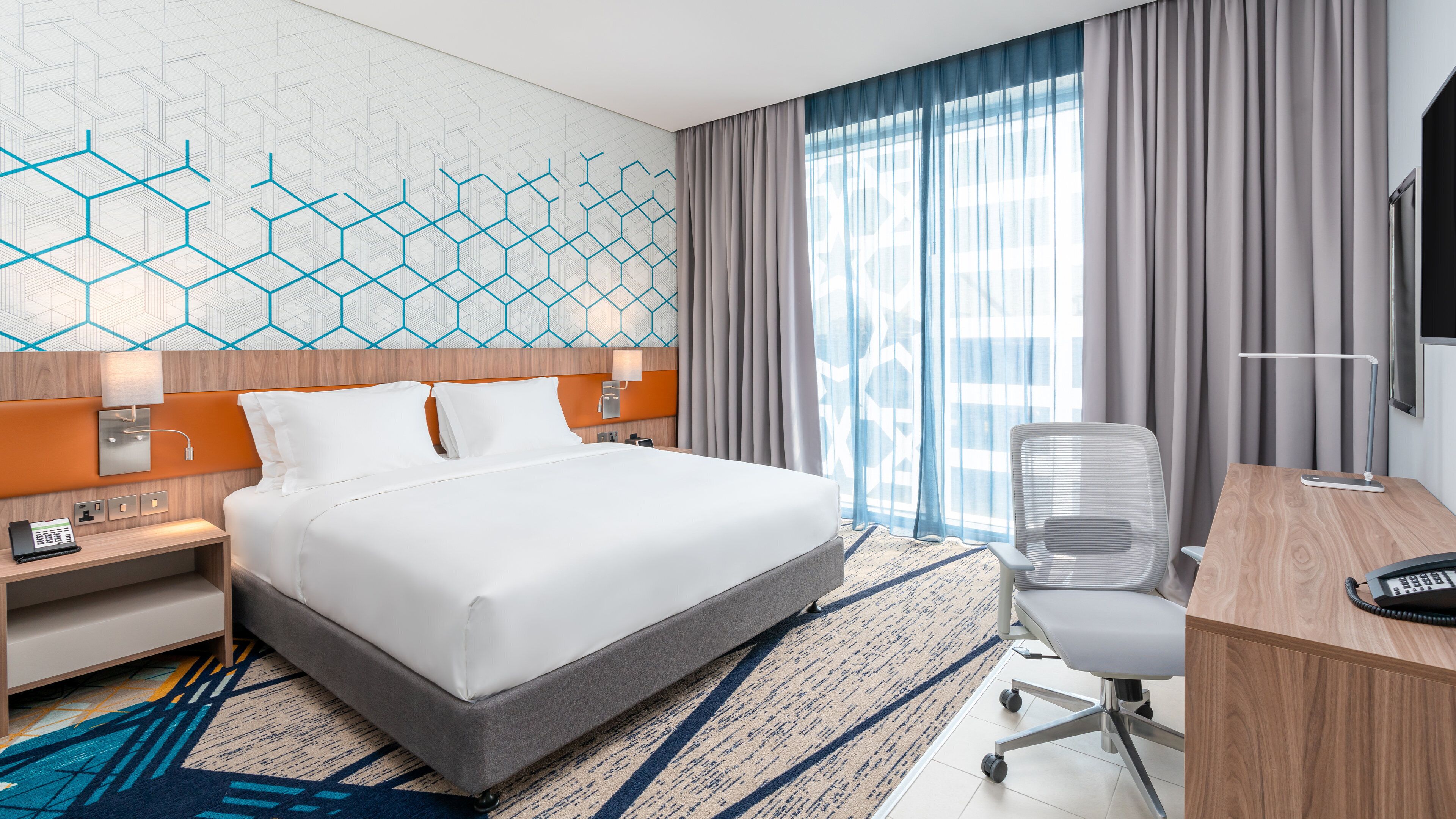 A hotel room with a large bed, a desk with a chair, a telephone, and a wall-mounted TV.
