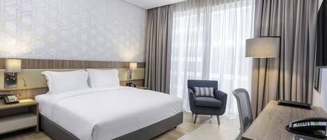 A modern hotel room with a large bed, a desk, a chair, and a lamp.