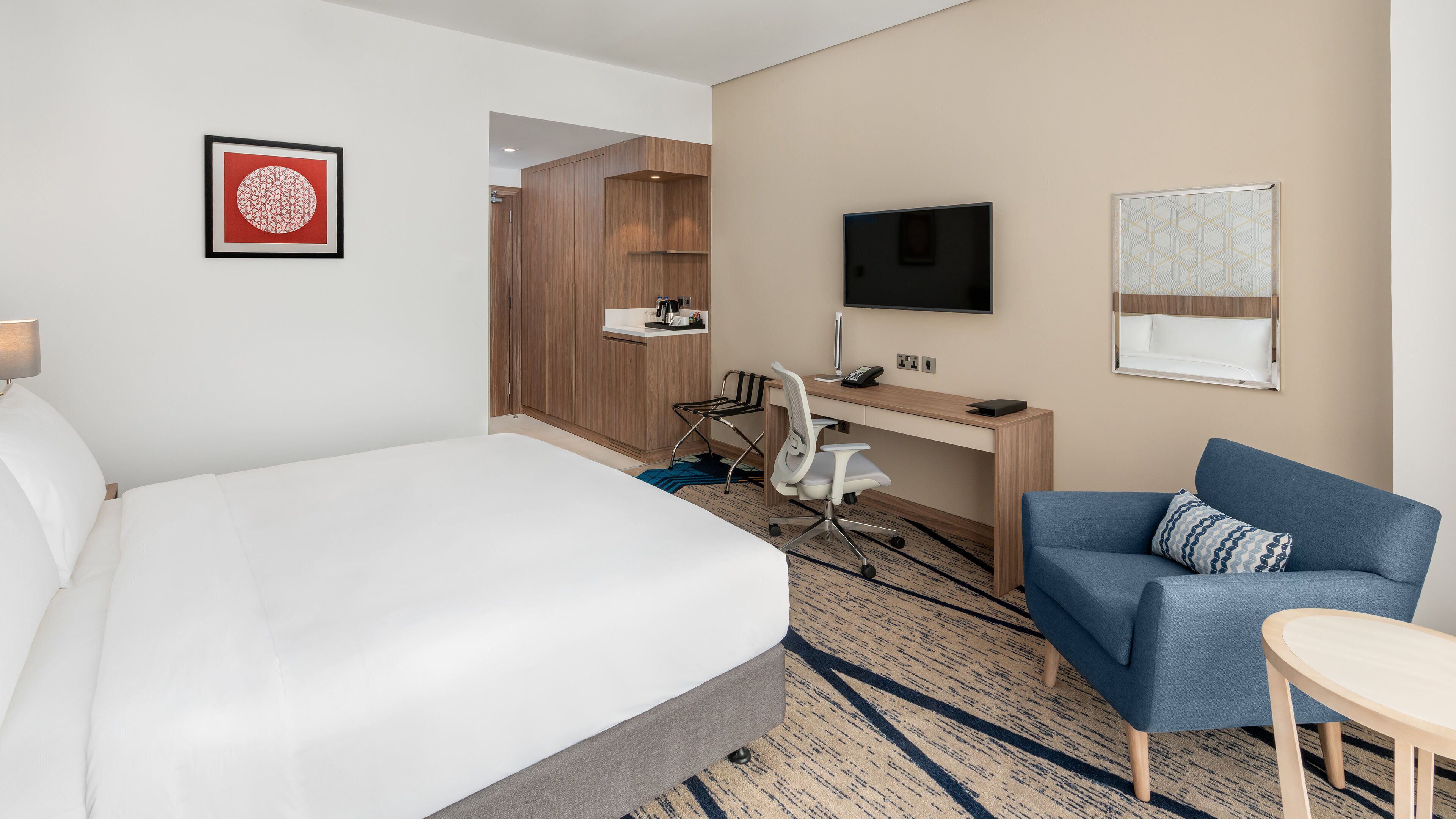 A hotel room with a bed, desk, chair, TV, and artwork.