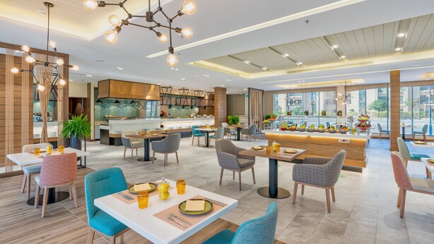 A modern restaurant with a central bar, multiple dining areas, and a kitchen visible in the background. - Holiday Inn & Suites Al Jubail by IHG (Al Jubail)