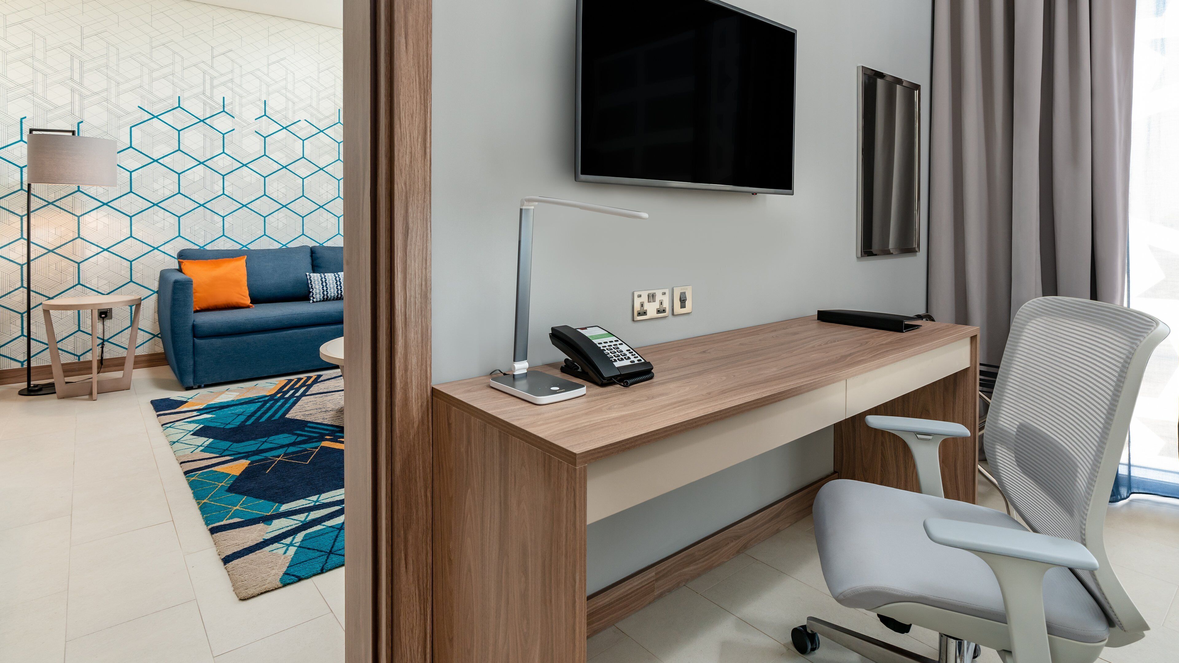 A modern hotel room with a wooden desk, a flat-screen TV, a sofa with a patterned rug, and a large window with curtains.