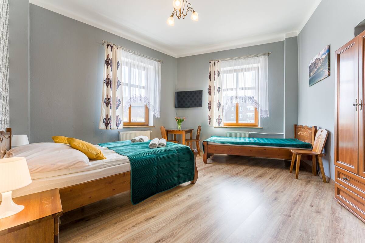 Quarto com duas camas, piso de madeira, escrivaninha, cadeira, televisão e guarda-roupa.