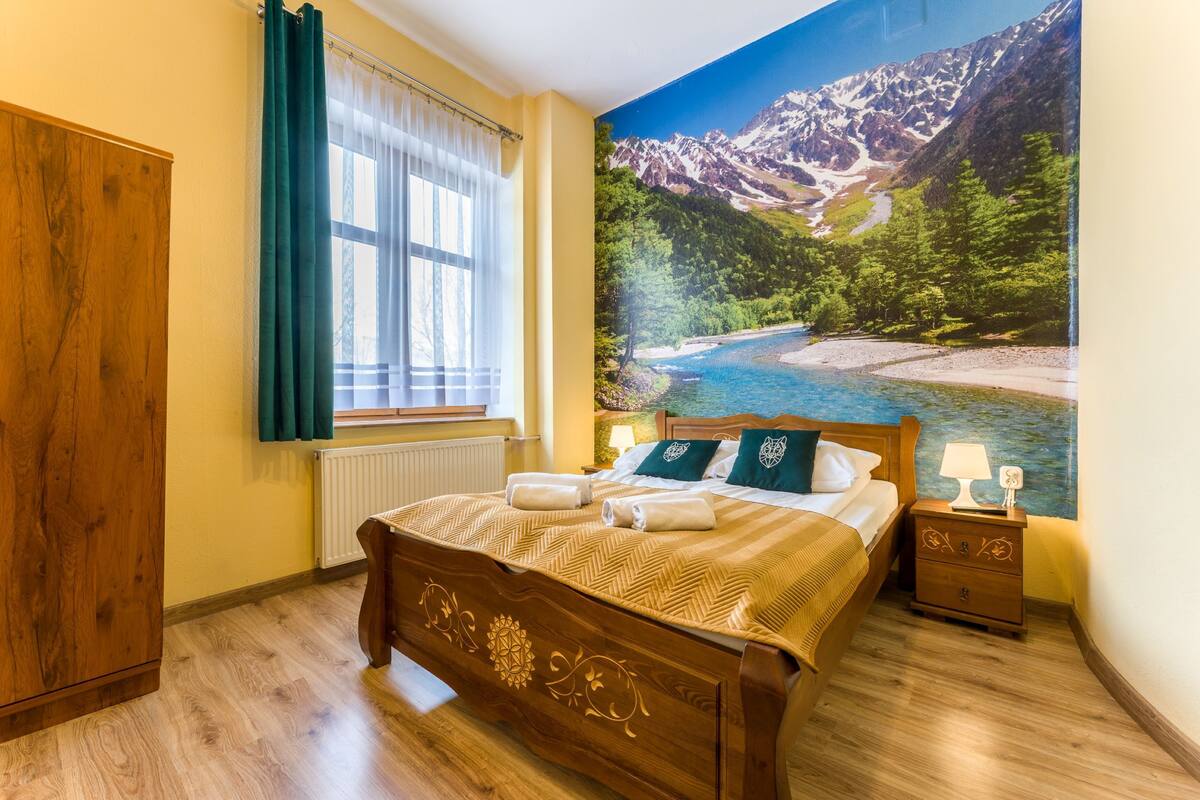 Quarto com cama de madeira, janela com cortinas, mesa de cabeceira com abajur e um mural de paisagem de montanhas.