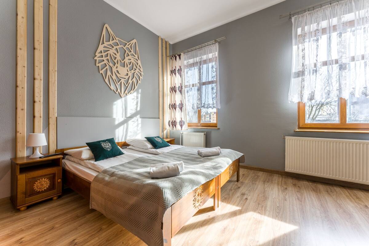 Quarto com cama, mesas de cabeceira, um detalhe decorativo na parede e janela com cortinas.