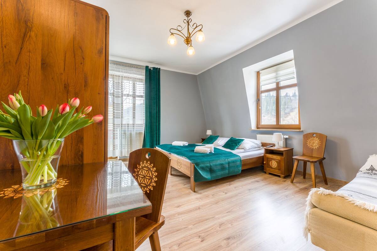 Quarto moderno com mesa de jantar de madeira, uma cama com cobertor verde, uma cadeira de madeira, mesa de cabeceira e janela com vista para árvores.