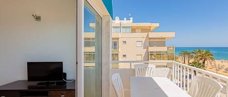 A balcony with a TV, a dining table, and chairs, overlooking a beach and buildings.