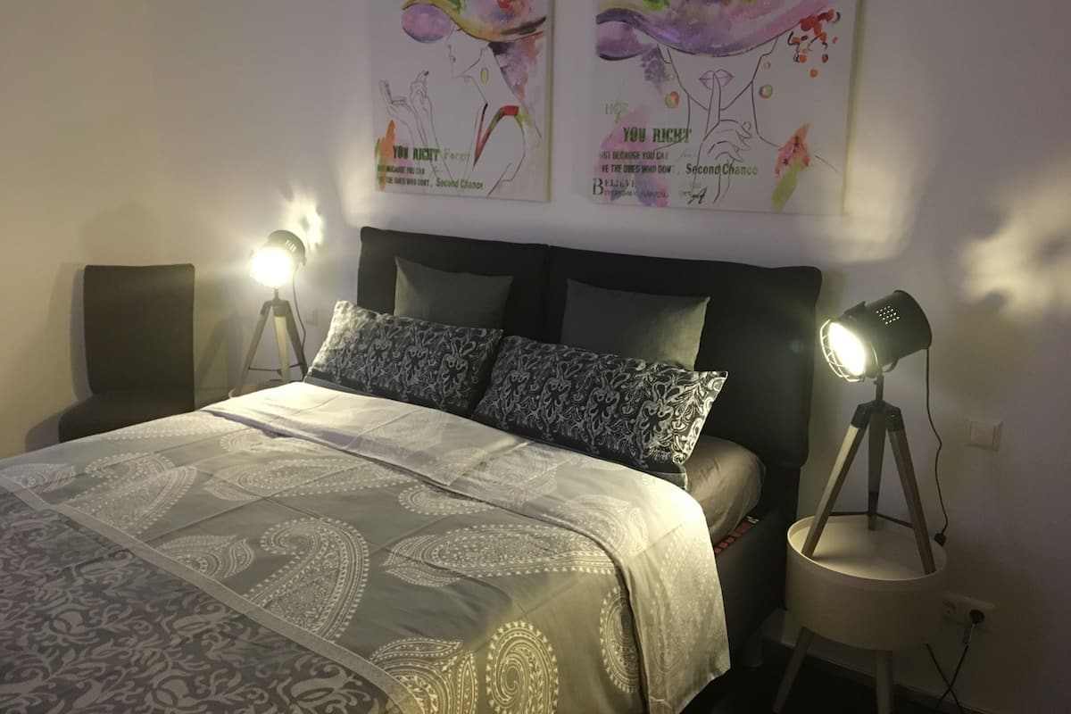 Uma cama bem arrumada com roupa de cama estampada, duas luminárias de cabeceira e duas obras de arte nas paredes acima da cama.