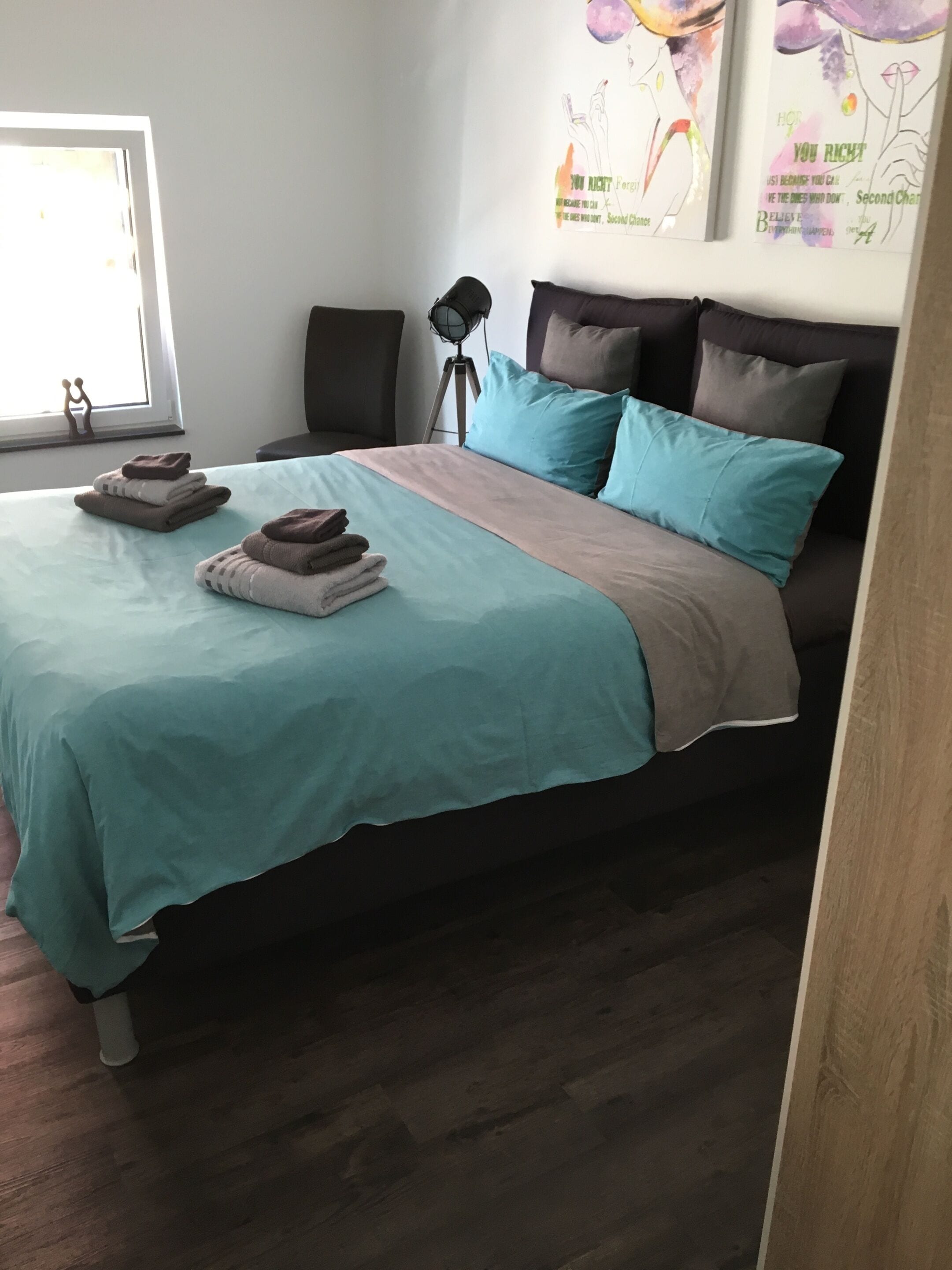 Un lit bien fait, avec une literie aux couleurs turquoise et beige, une lampe sur pied et une fenêtre donnant sur un beau paysage.