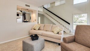 Ein Wohnzimmer mit einem beigefarbenen Sofa, einem grauen Hocker und einem braunen Sessel. Eine Treppe mit schwarzer Geländer führt zu einer oberen Ebene.