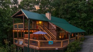 A two-story log cabin with a green roof, wooden deck, and a staircase leading to the upper level. - Free Tickets | Private Balcony, Grill, Game Room, Hot Tub, Great Location (Sevierville)