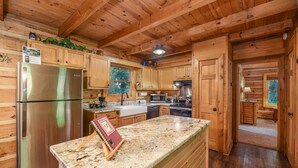 A wooden cabin kitchen with a stainless steel refrigerator, granite countertop, wooden cabinets, and a view of the outdoors through a window. - Free Tickets | Private Balcony, Grill, Game Room, Hot Tub, Great Location (Sevierville)