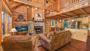 A log cabin interior with a stone fireplace, wooden furniture, and a kitchen area. - Free Tickets | Private Balcony, Grill, Game Room, Hot Tub, Great Location (Sevierville)