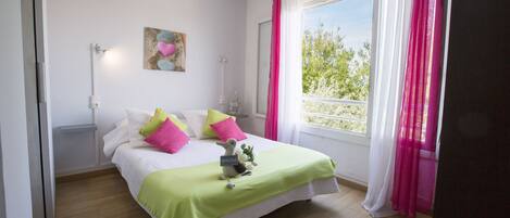 A bedroom with a bed, pillows, a bedside table, a lamp, a window with curtains, and a view of greenery outside.