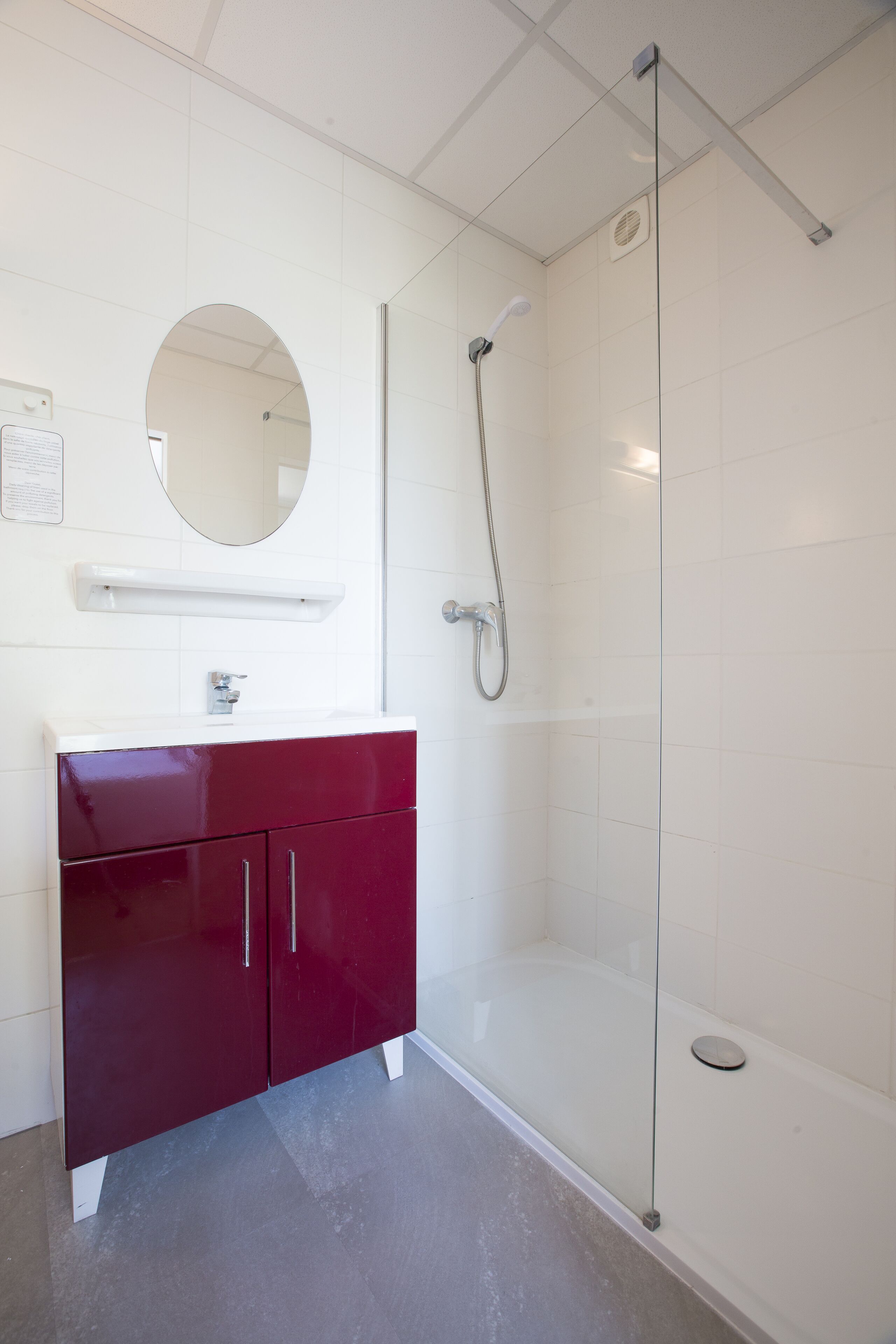 Ein modernes Badezimmer mit einer gläsernen Duschkabine, einem weißen Waschtisch mit rotem Unterschrank und einem runden Spiegel über dem Waschbecken.